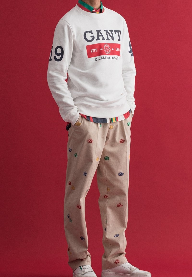 Sudadera blanca con el logo "GANT" en navy, combinada con pantalones beige que presentan patrones bordados coloridos, y zapatillas blancas.