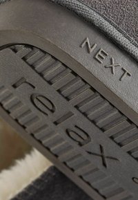 Chausson gris avec un dessus texturé, semelle en caoutchouc présentant les mots "relax" et "NEXT" embossés, doublure douce visible au bord.
