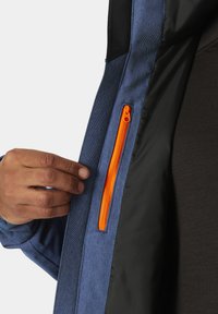 Giacca di jeans con una fodera interna nera, dotata di una tasca con zip arancione. Una mano afferra il tessuto della giacca, mettendo in risalto la sua texture e il design.