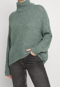 Przesadzony zielony sweter z dzianiny z wysokim kołnierzem typu stołowego, teksturowana tkanina, opadające ramiona i nierówne wykończenie, w połączeniu z ciemnymi dżinsami.