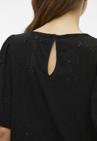 Svart blus med en nyckelhålsöppning på baksidan, texturerad tyg med mönstrade utskärningar och lösa, korta ärmar.