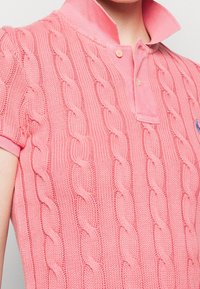 Różowy sweter polo z dzianiny o fakturze warkocza, z krótkimi rękawami i kołnierzykiem na guziki. Na piersi znajduje się małe niebieskie logo.