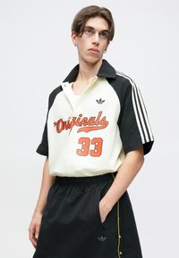 Kortermet skjorte i kremfarge og svart, med oransje "33" og "Originals" tekst. Svart shorts med sidespenner og gul kant.