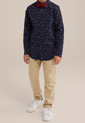 Niño con pantalones beige, zapatillas blancas, camisa azul marino con estampado navideño colorido y pajarita roja, de pie contra un fondo liso.