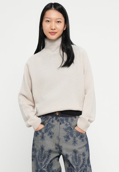 Mladá žena s dlhými čiernymi vlasmi má na sebe béžový rolák a modro-sivé vzorované nohavice, pričom má ruky v vreckách.