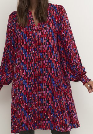 Donna che indossa un vestito a maniche lunghe con un motivo geometrico rosso, rosa, blu e nero e polsini arricciati sulle maniche.