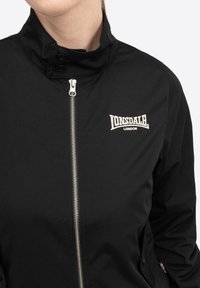 Schwarze Jacke mit Stehkragen, silberner Reißverschluss und aufgesticktem Logo auf der linken Brust. Glattes, langlebiges Material mit minimalistischem Design.