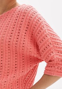 Koralowy sweter z dzianiny z ażurowym wzorem, szerokimi rękawami, luźnym krojem i teksturowanym prążkowaniem na ciele i rękawach.