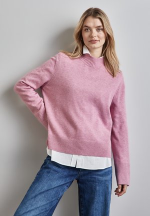 Pullover rosa a maglia con un alto collo a coste e maniche lunghe, indossato sopra una camicia bianca con colletto, abbinato a jeans blue.