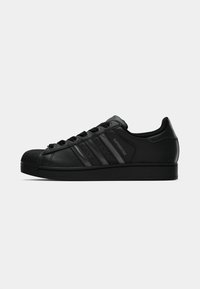 Black leather sneaker with a textured finish, rounded toe, and three tonal stripes. Features a rubber shell toe and flat laces.