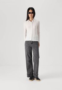 Long-sleeve, light beige button-up shirt, gray wide-leg jeans, and black sneakers with beige soles. Model wears dark sunglasses.