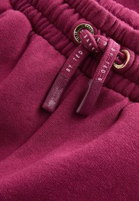 Bordeaux-housut, joissa on teksturoitu pinta, varustettuna kultaisilla yksityiskohdilla varustetulla nyörillä ja brändätyillä yksityiskohdilla. Elastinen vyötärö, joka mahdollistaa säädettävän istuvuuden.