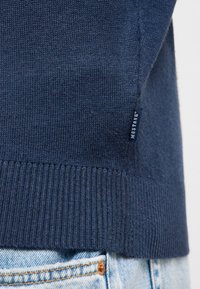 Ourlet de pull en maille bleu marine avec une étiquette de la marque Mustang au-dessus de la poche d'un jean denim bleu clair et rivet en cuivre.