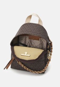 Brauner Canvas-Rucksack mit Logomuster, cremefarbenem Innenraum, goldener Kettenhenkel und Magnetverschluss. Mit einem oberen Griff zum Tragen.