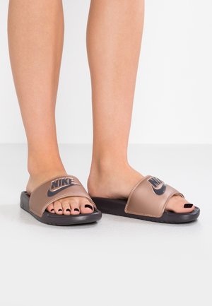 Pieds portant des sandales Nike slides en bronze avec semelles noires et ongles de pieds peints en noir sur un fond de sol gris clair.