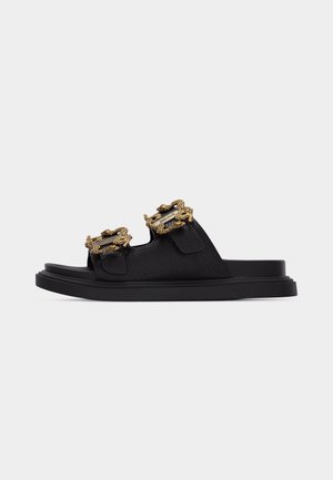 Sandalia negra tipo chancla con dos tiras texturizadas y hebillas ornamentadas en tono dorado sobre una suela gruesa.