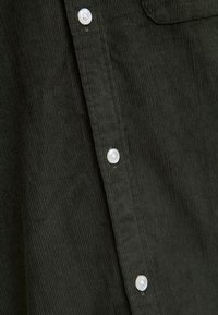 Mørkegrønn kordfløyelskjorte med vertikale riller, hvite knapper og en brystlomme; teksturert stoff gir en myk følelse og et strukturert utseende.