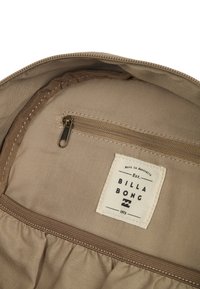 Interno beige di una borsa con una tasca con zip e un'etichetta chiara che riporta "BILLABONG" e l'anno di fondazione 1973. Texture liscia.
