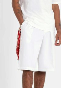 Shorts de sport blancs à coupe ample, dotés d'une taille élastique et d fentes latérales. Un tissu à motifs rouges dépasse de la poche.