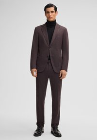 Abito da uomo in un ricco tessuto bordeaux con una vestibilità slim, caratterizzato da una giacca a due bottoni, rever a scollo e pantaloni con piega piatta. Indossato con un dolcevita nero.
