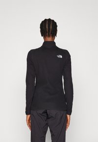 Zwart fleece pullover met een kraag, voorzien van een klein wit logo op de bovenrug, lange mouwen en een verstelbaar manchetdetail.