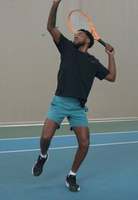 Jugador de tenis con una camiseta negra de manga corta y pantalones cortos color azul verdoso, sirviendo con una raqueta naranja en una cancha azul. Entorno interior, paredes texturizadas.