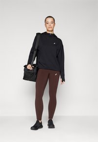 Black long-sleeve top with a high collar, brown leggings, and black shoes. The person holds a black bag with a textured surface and strap.