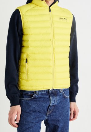 Mężczyzna ubrany w granatową koszulę z długim rękawem, żółtą pikowaną kamizelkę bez rękawów Red Bull Racing i niebieskie dżinsy na białym tle.