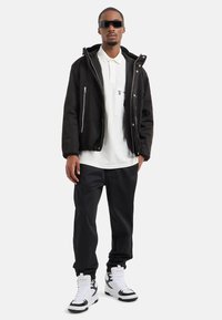 Giubbotto bomber nero con cerniere argentate, polo bianca, pantaloni scuri e sneaker alte in bianco e nero. Gli occhiali da sole completano il look.
