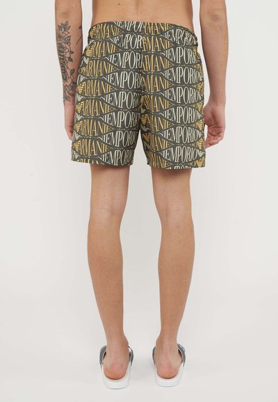 Uomo con pantaloncini Emporio Armani neri e oro con motivo a fantasia, in piedi a piedi nudi su un pavimento chiaro, vista posteriore e tatuaggi sulle gambe.