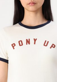 Κρεμ κοντομάνικο μπλουζάκι με ναυτικό μπλε γιακά, διακοσμημένο με τη φράση "PONY UP" σε κόκκινα αρθρωτά γράμματα στο στήθος.