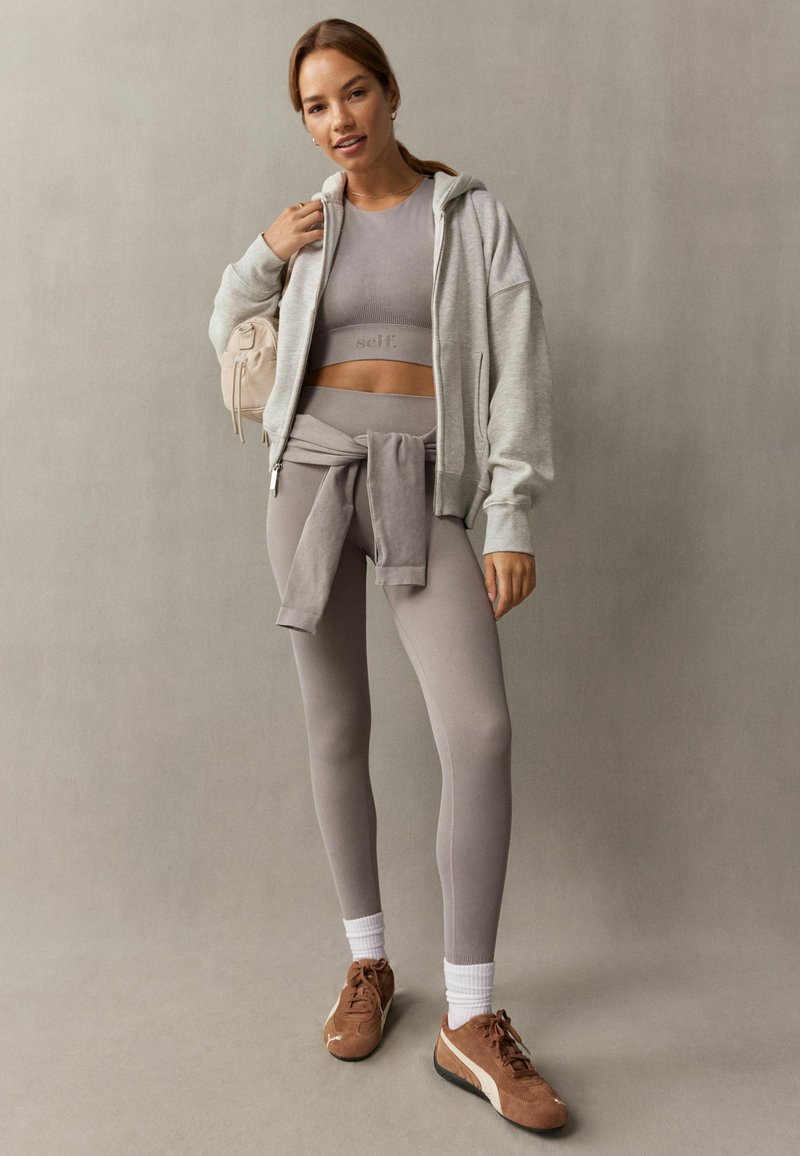 Jasnoszara bluza z zamkiem, dopasowany crop top i legginsy z wysokim stanem. Brązowe buty sportowe z białymi skarpetkami. Miękki, elastyczny materiał.