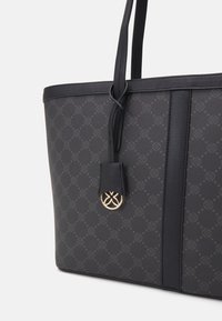 Sort tote bag med diskret mønsterdesign, to skulderstropper og et hængende lædermærke med et rundt guldemblem.