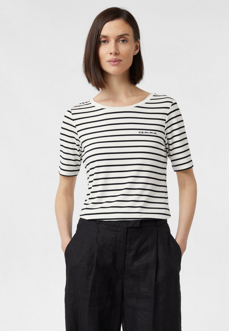 Gestreiftes T-Shirt in Weiß und Schwarz mit kurzen Ärmeln und Rundhalsausschnitt, das ein kleines "Komma"-Logo auf der Brust aufweist. Kombiniert mit dunklen Hosen.