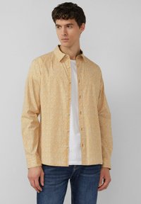 Lange mouwen overhemd met knopen in geel met een klein bloemenpatroon, gemaakt van lichtgewicht stof; gecombineerd met een wit t-shirt en blauwe spijkerbroek.