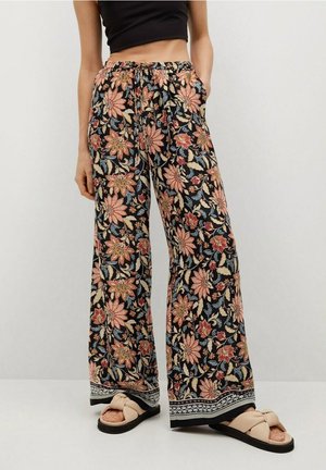 Kobieta ubrana w czarne, szerokie spodnie w kwiaty oraz beżowe sandały ze wstążkami, stojąca z jedną ręką w kieszeni.