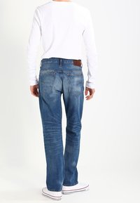 Džíny z denimu s volnějším střihem, vybledle modré barvy, s zadními kapsami, v kombinaci s bílým tričkem s dlouhým rukávem. Hnědá kožená záplata v pase.