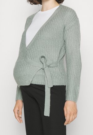 Osoba w ciąży ubrana w jasnoszary dziergany sweter typu wrap na białą koszulę i czarne spodnie, z swetrem zawiązanym z boku.