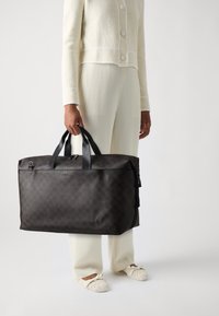 Dark brown checkered fabric duffel bag with black leather accents and handles, featuring a zip closure and a detachable shoulder strap.