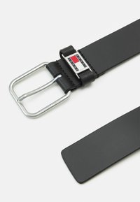 Zwarte leren riem met een zilveren metalen gesp. De gesp heeft een rechthoekig logo met rode, witte en blauwe accenten. Platte riemontwerp.