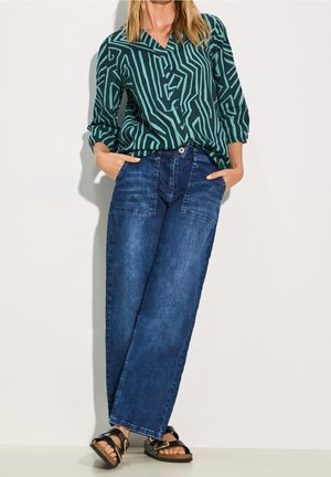 Frau trägt eine Bluse mit türkis-schwarzem geometrischem Muster, weite blaue Jeans mit aufgesetzten Taschen und schwarze Sandalen mit Schnallen.