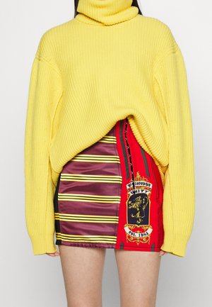 Femme portant un pull à col roulé jaune oversize et une mini-jupe rouge et violette avec des rayures jaunes et un blason à l'effigie d'un lion.