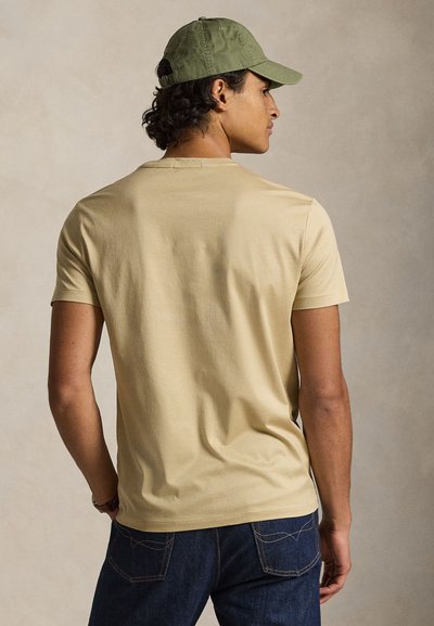 Κοντομάνικο, ανοιχτό καφέ T-shirt με στρογγυλή λαιμόκοψη, συνδυασμένο με σκούρο μπλε τζιν και σκούφο ελιάς, σε όψη από πίσω.