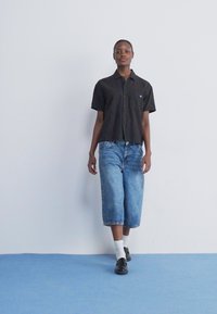 Svart randig kortärmad skjorta med knäppning, blå denimknä-långa shorts, vita strumpor och mörka skor mot en enfärgad bakgrund.