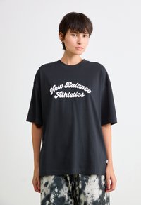 T-shirt en coton noir avec une coupe décontractée. Présente un texte blanc "New Balance Athletics" dans une police stylisée. Associe avec un pantalon tie-dye.