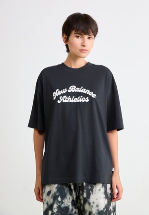 Camiseta de algodón negra con un corte relajado. Presenta texto blanco "New Balance Athletics" en una fuente estilizada. Combinada con pantalones de tie-dye.