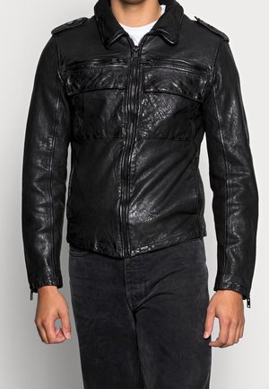 Mężczyzna noszący czarną, teksturowaną skórzaną kurtkę z zamkiem błyskawicznym i kieszeniami z klapami, w zestawieniu z czarnymi spodniami i białą koszulą pod spodem.