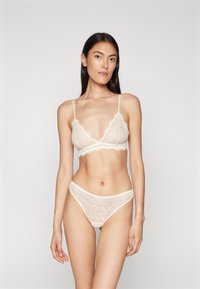 Lace-BHs-Set in Creme, bestehend aus einem Bralette mit gewellten Kanten und verstellbaren Trägern, passend zu einem Spitzen-Thong.
