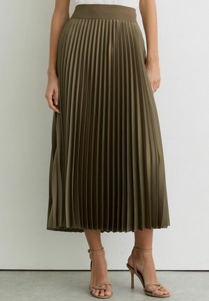 Frau trägt einen olivgrünen plissierten Midi-Rock und beige Sandalen mit hohen Absätzen, steht vor einer weißen Wand.