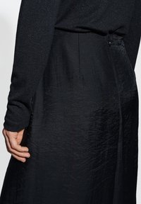 Robe noire avec une finition texturée, dotée de manches longues et d'un subtil éclat, mettant en valeur une fermeture éclair au dos et une taille cintrée.